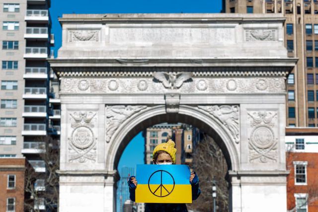 A young child in a yellow hat holds a blue and yellow poster painted in the Ukrainian flag colors with a peace sign, as the Washington Square Arch looms behind her.
